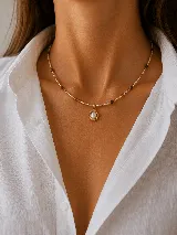Collar de cadena fina con cuentas pequeñas en tonos dorados, blancos y marrones oscuros, con un dije central ovalado enmarcado en dorado con una perla o piedra blanca en su interior.