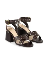 Sandalias de cuero color marrón chocolate, con taco ancho, tiras cruzadas en el empeine y tira al tobillo con hebilla metálica. Presentan tachas doradas decorativas en las tiras y en la sujeción del tobillo.