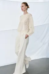 Camisa de algodón color crudo con cuello redondo y cierre de lazada en la espalda. Presenta mangas largas con puños, tablas y detalles de bordados y entredoses en el frente.