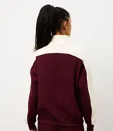 Buzo deportivo de corte oversized, con diseño bicolor en bordó y blanco. Presenta cuello alto con cierre de botones a presión, mangas largas con textura acanalada y puños elásticos.