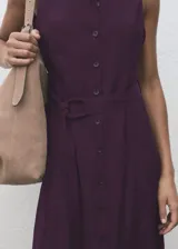 Vestido midi de corte evasé en color violeta oscuro, con cuello camisero, sin mangas y cierre frontal con botones. Incluye un cinturón ajustable a tono que marca la cintura.