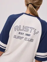 Remera bicolor Rusty de corte holgado y estilo deportivo, con cuerpo blanco y mangas raglán azules con doble raya blanca. Presenta estampado frontal con el texto 'RUSTY 85'.
