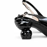 Zapato slingback de charol negro, con punta fina, taco bajo geométrico tipo joya y aplique metálico decorativo en la puntera. Cuenta con tira trasera ajustable con hebilla.