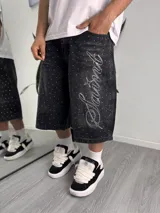 Bermuda de denim negro con diseño baggy, apliques de brillos en toda la prenda y gran bordado con texto en la pierna izquierda.