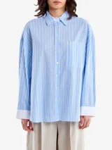 Camisa celeste y blanca a rayas verticales, de corte holgado, con cuello clásico, bolsillo en el pecho, mangas largas con puños blancos y cierre frontal con botones.