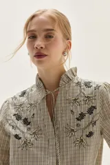 Blusa de algodón con estampado de rayas verticales en tonos crudo y negro. Presenta cuello mao con lazo para atar, bordados florales en negro y beige en la zona del pecho y hombros, y mangas largas con puños fruncidos elásticos.