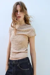 Top de punto color beige con cuello subido, manga corta y diseño texturizado con efecto arrugado. Presenta aplicaciones de perlas distribuidas en la parte frontal y las mangas.