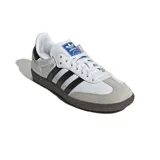 Championes Adidas modelo Samba OG, de diseño clásico con exterior de cuero blanco, tres tiras laterales negras, puntera de gamuza gris y suela de goma color marrón.