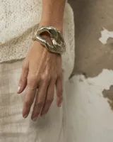 Brazalete abierto de diseño orgánico y voluminoso, con acabado metálico brillante. Su forma irregular y ergonómica permite un ajuste cómodo en la muñeca o antebrazo.