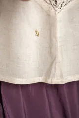 Blusa sin mangas de color beige, confeccionada en tela ligera con detalles de bordados florales frontales, encajes y cierre delantero con tiras para anudar.