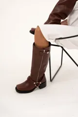 Bota de caña alta estilo biker confeccionada en cuero color chocolate. Presenta un cierre metálico lateral que recorre toda la caña, taco cuadrado y plataforma. Incluye un sistema de estribo y cadena metálica desmontable en la zona del tobillo, permitiendo diversas formas de uso.
