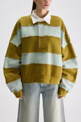 Buzo de estilo rugby con diseño oversize, confeccionado en bloques de color verde militar y celeste con costuras expuestas y deshilachadas. Cuenta con cuello camisero blanco de popelina y cartera de botones frontal oculta, además de mangas largas y puños acanalados.