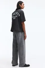 Remera boxy de algodón gris oscuro, con corte oversized, hombros caídos, cuello redondo ribeteado y estampa pequeña en el pecho con texto en blanco.