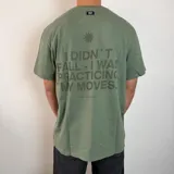 Remera de algodón color verde oliva con estampa gráfica en la espalda que incluye un sol y la frase 'I didn't fall - I was practicing my moves'. Presenta un corte regular y etiqueta pequeña en la parte superior trasera.