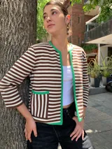Chaqueta corta tejida a rayas horizontales marrones y blancas, con cuello redondo y ribete verde. Tiene bolsillos frontales y mangas 3/4.