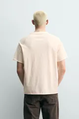 Camiseta relaxed fit de algodón color beige claro, con cuello redondo y mangas raglán cortas. Cuenta con estampado frontal en tono azul con efecto desgastado que incluye texto y el número 87.