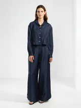 Camisa de manga larga color azul marino, confeccionada en lino italiano con hilo metalizado que le da un sutil brillo. Presenta cuello clásico y botones frontales, con un detalle de broche o dije en forma de flor en el cuello.