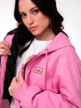 Chaqueta corta de lona color rosa, con capucha ajustable, cierre frontal y forro interior acolchado negro. Presenta un parche con el logo de la marca en el pecho.