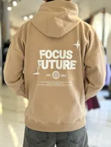 Canguro oversize tostado con capucha y bolsillo tipo canguro, confeccionado con diseño holgado. En la espalda presenta un estampado en color blanco con las palabras "FOCUS FUTURE", gráficos de estrellas y un diseño de globo terráqueo acompañado del texto "EST. 2021".