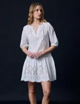 Vestido corto blanco de algodón con bordado calado tipo broderie, cuello redondo, escote en V con lazo y borlas, y mangas tres cuartos con puño.