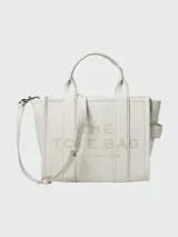Cartera tipo tote bag Marc Jacobs modelo The Leather Small Tote Bag, color negro, confeccionada en cuero graneado con el texto 'THE TOTE BAG' grabado en relieve al frente. Cuenta con asas superiores, correa ajustable y desmontable, y herrajes metálicos dorados.