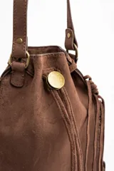 Cartera tipo bucket color marrón, confeccionada en material sintético con acabado gamuzado. Presenta flecos largos en los laterales y la base, cierre mediante cordón ajustable y correa larga regulable para llevar al hombro o cruzada.