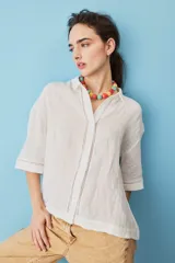 Camisa blanca de lino, con mangas anchas a tres cuartos, cuello clásico, cierre frontal con botones y detalles de calados verticales en la parte central y en los puños.