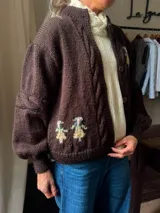 Cardigan de punto color marrón, con diseño de ochos y bordados artesanales de figuras en la parte inferior, cuello alto y cierre frontal con botones.