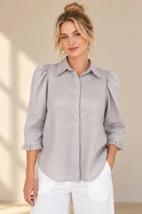 Camisa de lino color blanca, con cuello clásico, mangas tres cuartos abullonadas en los hombros y terminación con volados en los puños, cierre frontal con botones ocultos.