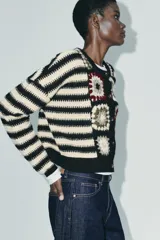 Chaqueta de punto con cuello redondo y manga larga, que combina un diseño de rayas horizontales en blanco y negro con paneles frontales decorados con motivos de crochet en tonos tierra y granate. Presenta cierre frontal con botones.