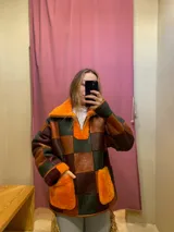 Buzo de cuero de oveja y diseño patchwork en tonos anaranjado, marrón y verde, con cuello alto, cierre frontal parcial y bolsillos frontales de corderito.