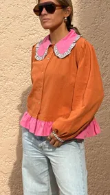 Camisa de manga larga color naranja con cuello grande en contraste color rosa y detalle de volados en el borde. Presenta un diseño con botones frontales y un detalle de tela rosa en la parte inferior.