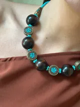 Collar corto con cuentas de madera color marrón oscuro, cuentas tibetanas con incrustaciones de color turquesa y detalles en metal color bronce. Cierre con cadena de acero quirúrgico y gancho para regular el largo.