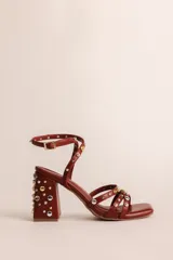 Sandalias de tacón ancho de 7 cm, color bordó, con diseño de tiras cruzadas en el empeine y pulsera al tobillo con cierre de hebilla. El modelo está decorado con tachas metálicas plateadas y doradas distribuidas por todas las tiras y el tacón.