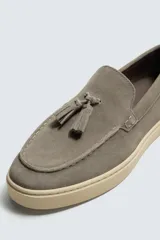 Mocasín casual de piel con acabado de serraje en color gris, con detalle de borlas en el empeine y suela plana a contraste en color beige.