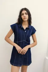 Vestido corto camisero de jean azul oscuro con cuello clásico, cierre frontal con botones y mangas cortas. Presenta costuras decorativas en color blanco.
