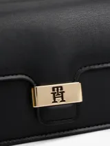 Cartera tipo bandolera de color negro, con acabado liso y solapa frontal. Presenta herrajes metálicos dorados, incluyendo un cierre con el monograma TH y un logo de Tommy Hilfiger en bajo relieve.