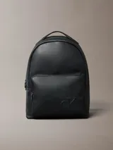 Mochila Calvin Klein de poliuretano color negro, de diseño minimalista con silueta redondeada. Cuenta con un compartimento principal con cierre, bolsillo frontal externo con cierre y el monograma CK grabado. Incluye asa superior y correas acolchadas ajustables.