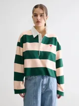 Remera estilo rugby oversize de manga larga y largo cropped, con diseño a rayas horizontales en verde oscuro y rosado. Posee cuello camisero blanco en contraste, cartera de botones, pequeño bordado en forma de corazón en el pecho y terminación desflecada en el ruedo.