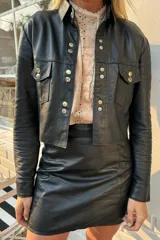 Camisa de cuero negro con corte a la cintura, cuello clásico y cierre frontal con botones metálicos en tonos dorados y plateados. Presenta dos bolsillos frontales con solapa y botones a juego.