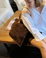 Bolso tipo tote de gamuza color marrón, con costuras visibles y bolsillo frontal externo. Cuenta con doble correa de cuero en tono natural para llevar al hombro o cruzado.