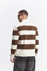 Polo de manga larga con diseño de rayas horizontales anchas en color marrón y crema. Presenta cuello camisero blanco con cartera de botones y estampado frontal con texto decorativo y logo de la marca.