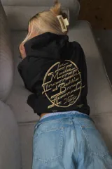 Canguro negro de corte oversize con capucha y bolsillo frontal. Presenta un diseño gráfico circular estampado en color beige en la espalda con tipografía estilizada y detalles de texto en la parte inferior.