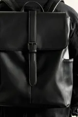 Mochila de diseño minimalista en color negro, confeccionada en material sintético con acabado liso. Presenta una solapa frontal con cierre de hebilla decorativa y sistema de imán, compartimento principal con cremallera, bolsillos laterales externos, asa superior de mano y correas de hombro ajustables y acolchadas.