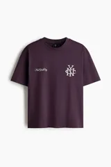 Remera de algodón color bordó con cuello redondo acanalado, mangas cortas y hombros caídos. Presenta un estampado frontal con el texto 'New York City' y un monograma entrelazado en color blanco.