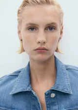 Chaqueta de jean azul de corte crop, con cuello camisero, cierre frontal de botones metálicos, bolsillos con solapa en el pecho y mangas largas con puños abotonados.