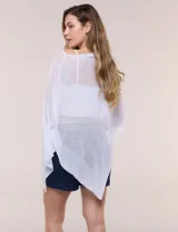 Poncho liviano de tejido de punto fino calado, color blanco, con silueta asimétrica envolvente, escote bote drapeado y mangas integradas estilo capa.