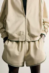 Bermuda de corte relajado confeccionada en tejido de algodón, color beige con vivos en contraste color verde oscuro. Cuenta con cintura elástica ajustable con cordón, bolsillos frontales y bolsillos traseros de plastrón.