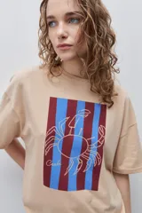 Remera blanca de calce relajado y silueta amplia, con cuello redondo, mangas cortas y estampa gráfica frontal de un cangrejo sobre franjas en tonos rosado y rojo.