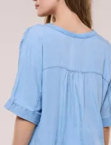Blusa estilo denim de lyocell celeste, de silueta amplia y suelta, con escote en V, cartera frontal de botones y mangas cortas japonesas de hombros caídos. Cuenta con ruedo asimétrico y posibilidad de anudar al frente.